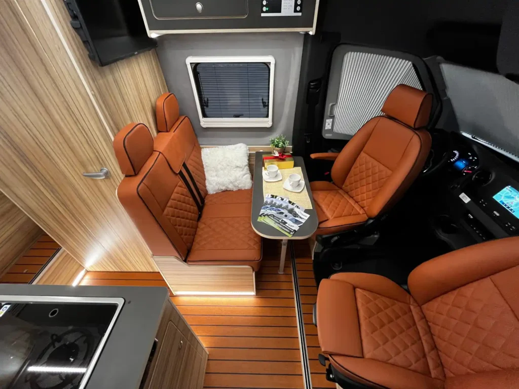 Wnętrze kampera z luksusowymi skórzanymi fotelami w kolorze brązowym, rozkładanym stolikiem i widoczną częścią kuchenną