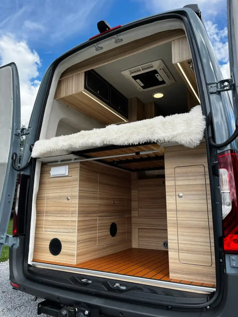 Otwarte tylne drzwi kampera ukazujące eleganckie drewniane wykończenie wnętrza, wbudowane szafki i puszystą narzutę na łóżku